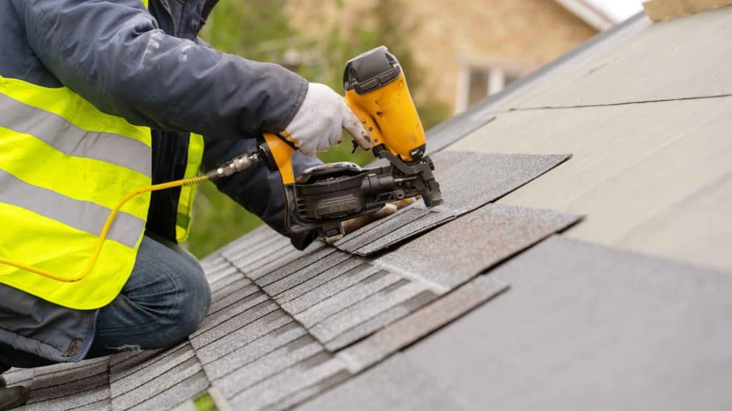 A person using a nail gun to apply composite shingles to a roof.